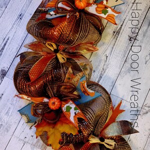 May include: A brown, orange, and gold fall wreath with a burlap bow and small pumpkins. The wreath is made of mesh and has a variety of fall-themed decorations. The text "Happy Door Wreaths" is visible in the background.