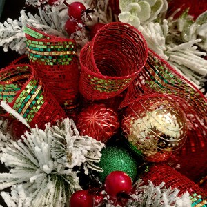 May include: A Christmas wreath featuring red and green mesh ribbon, red berries, and gold and red baubles. The wreath is decorated with white faux snow.