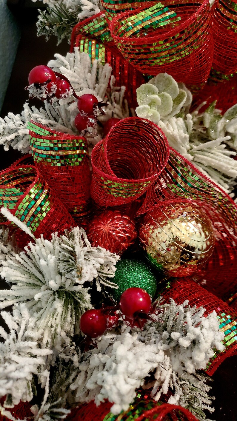 May include: A Christmas wreath featuring red and green mesh ribbon, red berries, and gold and red baubles. The wreath is decorated with white faux snow.