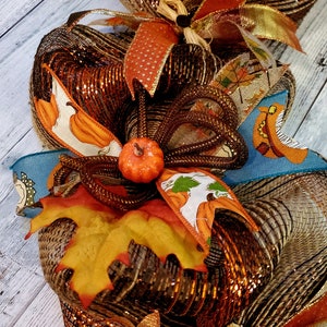May include: A fall-themed wreath with brown, orange, and gold mesh, accented with a small orange pumpkin, a brown bow, and a fall leaf.