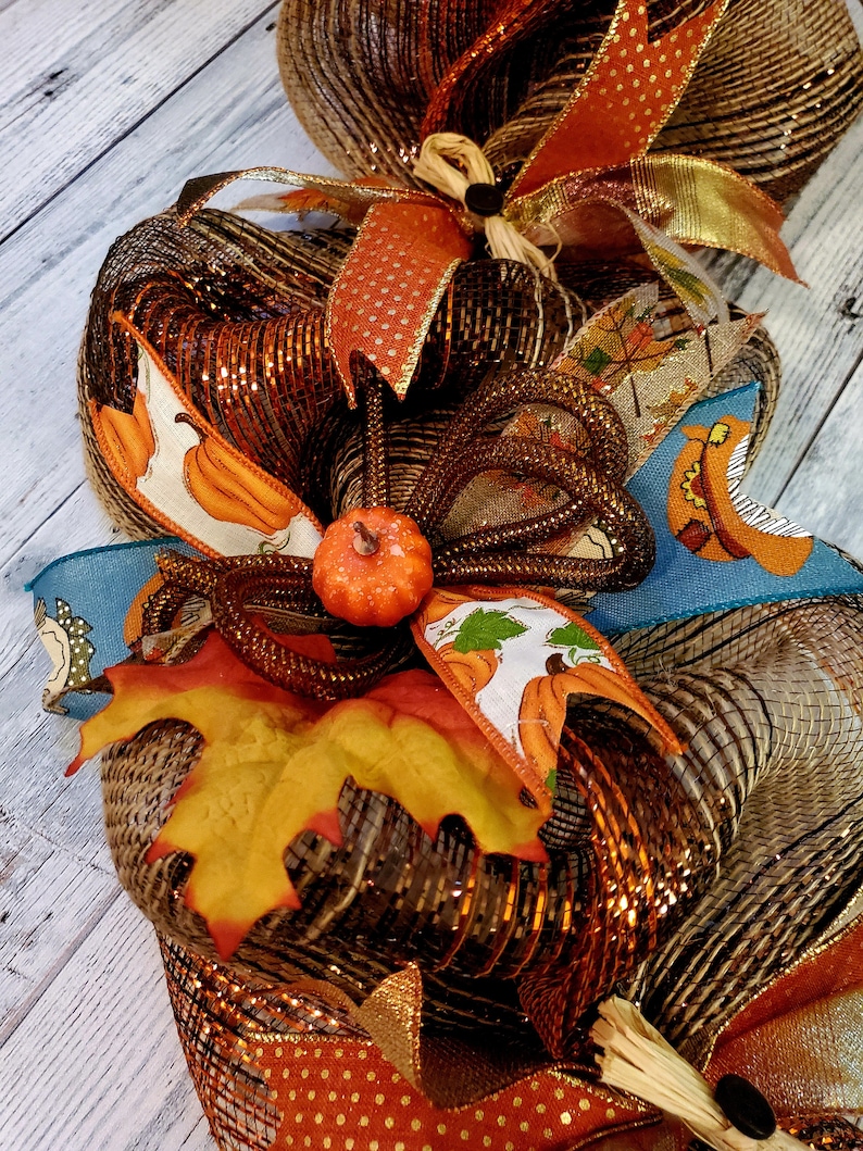 May include: A fall-themed wreath with brown, orange, and gold mesh, accented with a small orange pumpkin, a brown bow, and a fall leaf.
