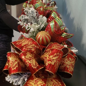 May include: A red and gold Christmas tree topper with a large bow and a glittery red and green bauble. The bow is made of a red ribbon with gold holly leaves and berries.