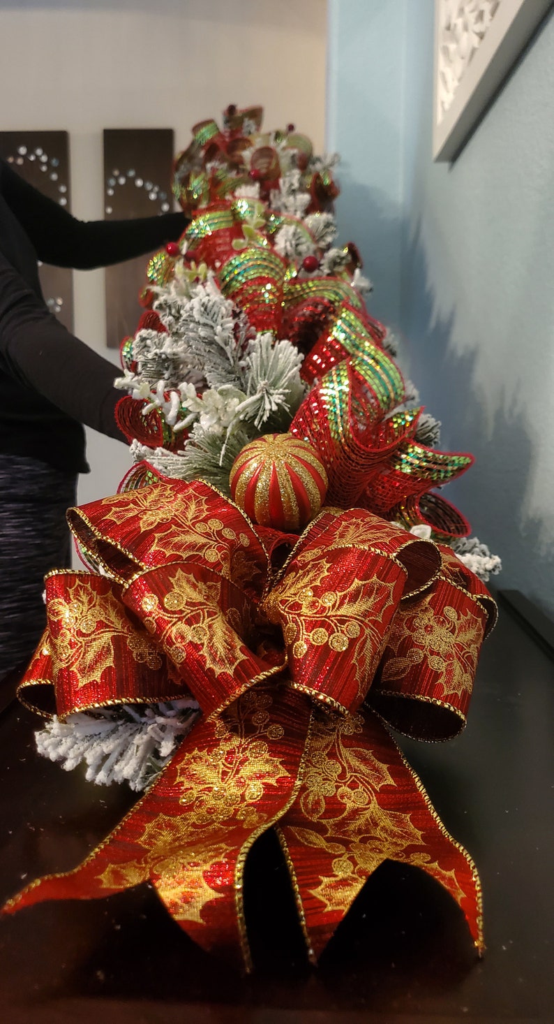 May include: A red and gold Christmas tree topper with a large bow and a glittery red and green bauble. The bow is made of a red ribbon with gold holly leaves and berries.