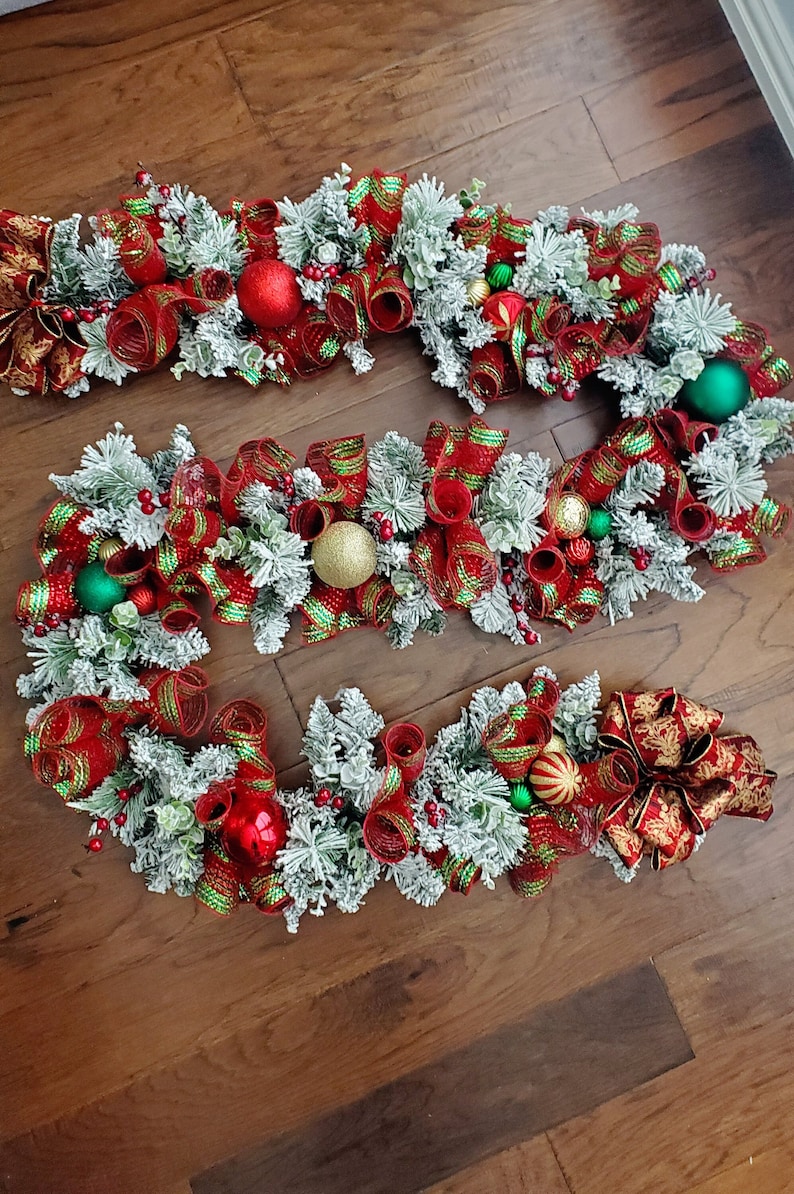May include: A red and green Christmas garland with a gold bow. The garland is decorated with red and gold baubles, green pine sprigs, and white faux snow.