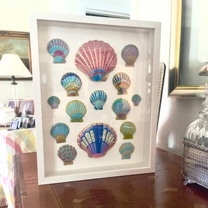 May include: A white framed art piece featuring colorful seashell designs. The shells are in various sizes and colors, including pink, blue, and gold. The artwork is displayed on a wooden surface, with a lamp and other decorative items in the background.