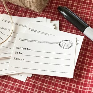 以下が含まれることがあります： スプーンのイラストと書き込み用の線が入った白い長方形のラベルの束。ラベルの隣には、ひもと黒いSharpieペンがあります。ラベルは赤と白のチェック柄の生地の上にあります。