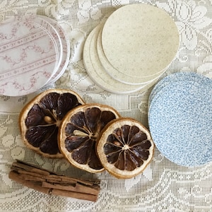 May include: A set of four dried orange slices, a cinnamon stick, and two sets of decorative paper coasters. One set of coasters is white with a pink floral pattern, and the other set is blue with a white floral pattern.