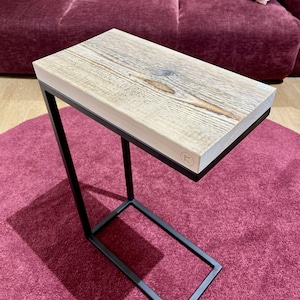 Op de afbeelding: Een bijzettafel met een rechthoekig, licht houten blad en een zwart metalen frame. De houtnerf is zichtbaar op het tafelblad. De tafel is ontworpen met een C-vorm, geschikt om naast een bank of stoel te plaatsen. De tafel staat op een rood tapijt.
