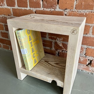 Puede incluir: Una mesa auxiliar de madera clara con un estante, que contiene un libro con una cubierta amarilla y verde. La mesa tiene un diseño de marco cuadrado y un pequeño logotipo redondo en el lateral. La mesa está hecha de madera clara.
