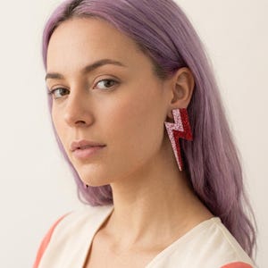 May include: A lightning bolt shaped earring featuring a dual-tone design with a glittery light pink section on the left and a glittery dark red section on the right. The earring has a sharp, angular silhouette and a textured, sparkling finish.
