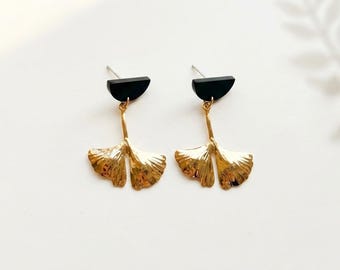 Puede incluir: Un par de pendientes colgantes con un dije de hoja de ginkgo de metal dorado suspendido de un componente de perno semicircular negro. Los pendientes tienen un cierre de presión y un acabado pulido, mostrando el diseño de hoja en abanico.