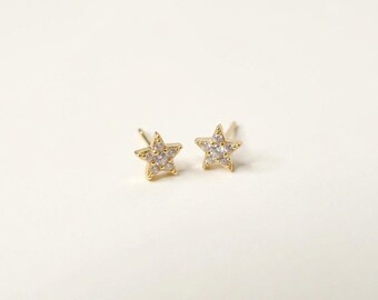 May include: A pair of gold-toned star-shaped stud earrings. Each earring features a five-pointed star design with a central cluster of small, clear, sparkling stones embedded within the metal frame, complete with a post backing for secure attachment.