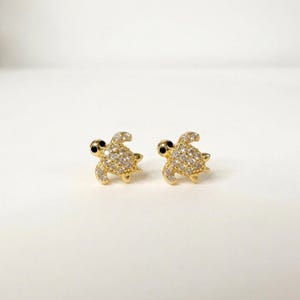 Puede incluir: Un par de pendientes de botón en forma de tortugas marinas doradas. Cada tortuga presenta un caparazón incrustado con pequeñas piedras claras y brillantes, y una piedra negra para el ojo sobre un acabado de metal color oro.