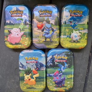 May include: Five rectangular metal Pokémon Trading Card Game tins featuring colorful illustrations of various Pokémon characters in outdoor landscapes. Each tin displays the Pokémon Trading Card Game logo at the top with mountain scenery.