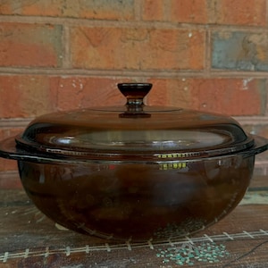 May include: A round, amber-colored glass casserole dish with a matching lid. The dish features a smooth, translucent finish and a central knob handle on the lid. The design is simple and functional, showcasing the deep brown hue of the glass material.