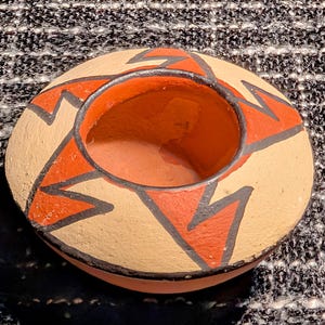 May include: A small, round, terracotta-colored pottery bowl with a wide opening. The exterior is tan with a repeating pattern of red and black geometric shapes. The interior is a vibrant orange. The bowl is likely handmade.