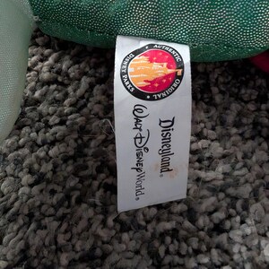 May include: A white tag with the words "Disneyland" and "Walt Disney World" printed in black. The tag also features a circular logo with the words "Authentic Original" and a colorful illustration. The tag is attached to a green plush toy.