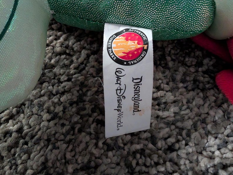 May include: A white tag with the words "Disneyland" and "Walt Disney World" printed in black. The tag also features a circular logo with the words "Authentic Original" and a colorful illustration. The tag is attached to a green plush toy.