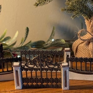May include: Miniature decorative black metal gates with white pillars and a detailed design. The gates are set on a wooden surface, with small evergreen trees in the background, creating a charming, rustic scene.