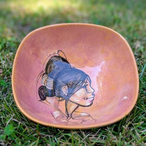 May include: A handmade, square-shaped ceramic bowl with a hand-painted illustration of a woman's profile. The bowl has a warm, reddish-brown hue with a glossy finish. The woman's portrait is rendered in black and white with yellow accents.