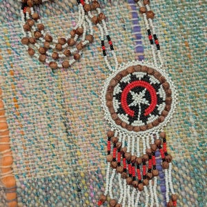 May include: Handmade beaded necklace featuring a central circular pendant with a red, black, and white design. The necklace includes multiple strands of brown beads and white seed beads, with red and brown accents. The pendant has a tree-like design.