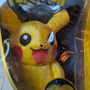 May include: A yellow Pikachu plush toy with red cheeks, black eyes, and a red tongue, packaged in a clear plastic bag. The bag has the "Pokémon" logo in blue and yellow. A circular emblem with the number "25" is visible.