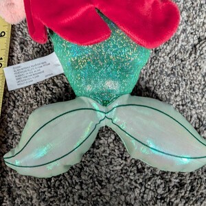 May include: A plush mermaid doll with red hair, a green sequined top, and a shimmering green tail. The doll has pink accents and is made of soft materials. The doll is a Disney product.