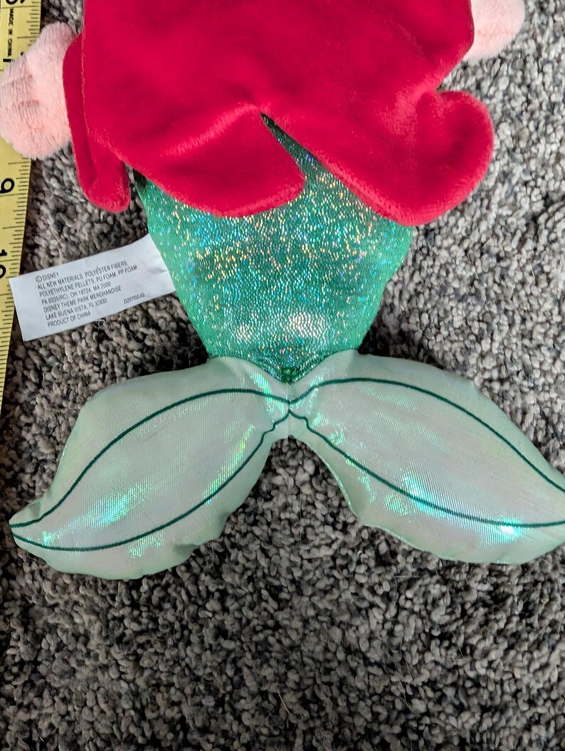 May include: A plush mermaid doll with red hair, a green sequined top, and a shimmering green tail. The doll has pink accents and is made of soft materials. The doll is a Disney product.