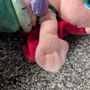 May include: A plush doll of a character with pink skin, red hair, and a green tail. The doll has a light pink body, a purple top, and a green tail. The doll is lying on a gray, textured surface. The doll has a Disneyland tag.