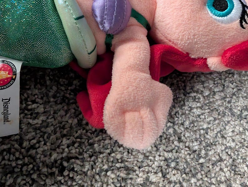 May include: A plush doll of a character with pink skin, red hair, and a green tail. The doll has a light pink body, a purple top, and a green tail. The doll is lying on a gray, textured surface. The doll has a Disneyland tag.