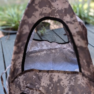 May include: A small, tent-like structure with a digital camouflage pattern in shades of gray and tan. The tent has a black mesh window and a black trim. The structure is designed to provide shelter.