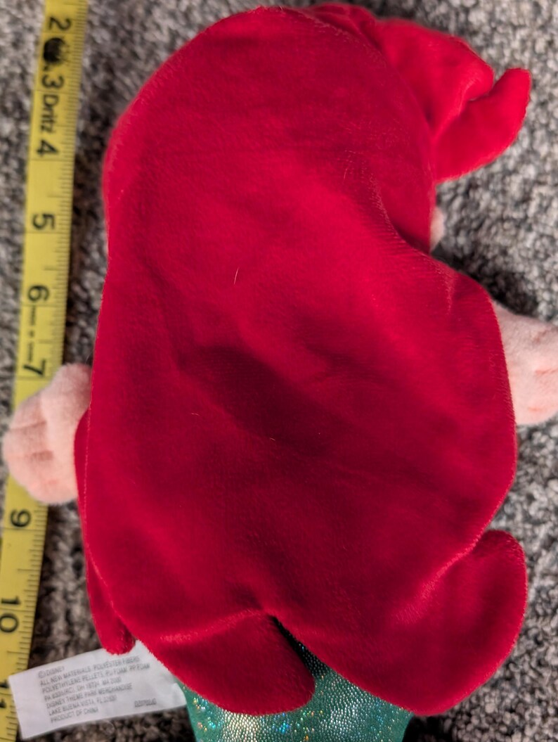 May include: A plush doll featuring a red velvet hair and a green sequined tail. The doll's hair is styled to the side. The doll is approximately 6 inches tall.