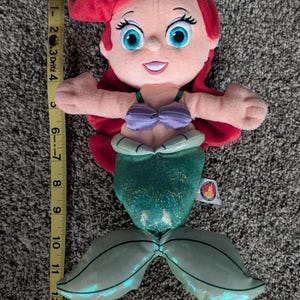 May include: A plush doll of a mermaid with red hair, blue eyes, and a purple top. The mermaid's tail is green and measures approximately 12 inches long. The doll has a smiling expression and outstretched arms.