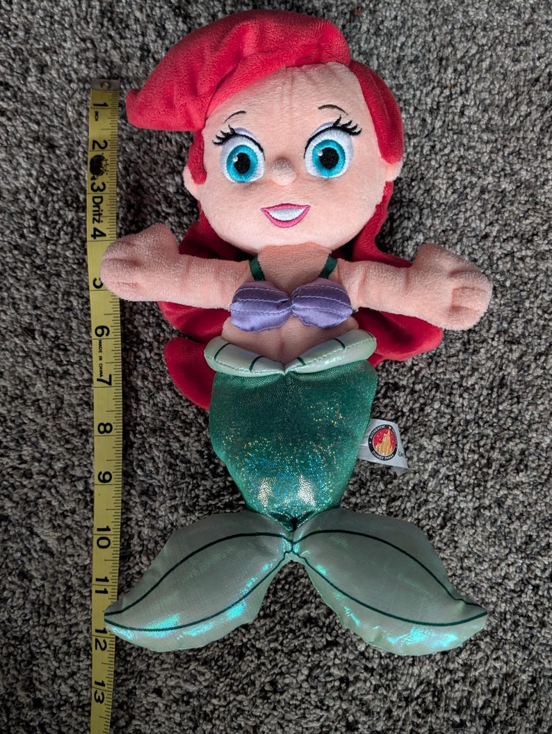 May include: A plush doll of a mermaid with red hair, blue eyes, and a purple top. The mermaid's tail is green and measures approximately 12 inches long. The doll has a smiling expression and outstretched arms.