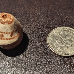May include: A small, white ceramic bead with brown painted designs. The bead has a hole in the top and is about the size of a US quarter.