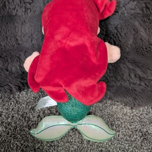 May include: A plush mermaid doll with bright red hair, a green sequined tail, and iridescent fins. The doll's arms and face are a light pink color. The doll is lying on a gray surface.