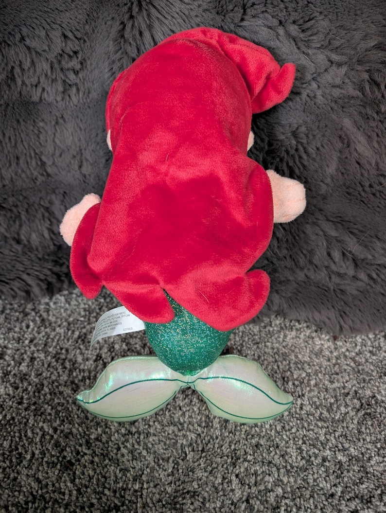 May include: A plush mermaid doll with bright red hair, a green sequined tail, and iridescent fins. The doll's arms and face are a light pink color. The doll is lying on a gray surface.