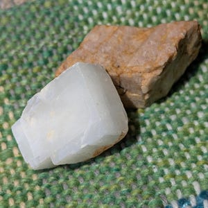 May include: Two raw gemstones, one a pale, translucent white, the other a rough, tan-colored stone. The white stone is in the foreground, with the tan stone behind it. The background is a green, blue, and white woven fabric.