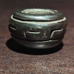 May include: A dark, round bead with a cylindrical shape and a central hole. The bead is black with carved, geometric designs around its circumference. The bead is likely made of wood or a similar material.