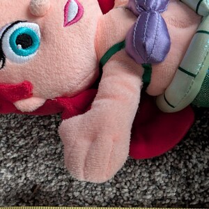 May include: A plush doll featuring a character with pink skin, red hair, and a teal eye. The doll has a purple bow and a green and silver tail. The doll is lying on a textured surface.
