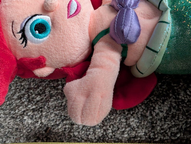 May include: A plush doll featuring a character with pink skin, red hair, and a teal eye. The doll has a purple bow and a green and silver tail. The doll is lying on a textured surface.