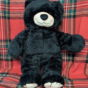 May include: A plush black teddy bear with a cream-colored snout and foot pads. The bear is standing on a red and green plaid blanket. The bear has a friendly expression.