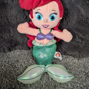 May include: A plush doll of a mermaid with red hair, blue eyes, and a purple seashell top. The mermaid has a green, glittery tail and a tag attached. The doll is designed to resemble a popular animated character.