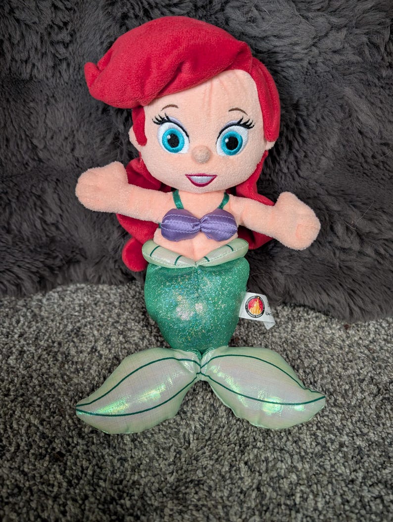 May include: A plush doll of a mermaid with red hair, blue eyes, and a purple seashell top. The mermaid has a green, glittery tail and a tag attached. The doll is designed to resemble a popular animated character.