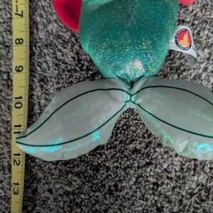 May include: A plush doll of a mermaid with a green sequined tail and bodice. The doll has red hair and a purple top. The tail measures approximately 6 inches from the top of the bodice to the end of the tail.
