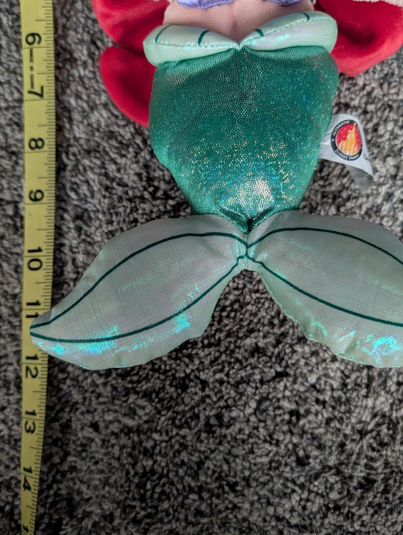May include: A plush doll of a mermaid with a green sequined tail and bodice. The doll has red hair and a purple top. The tail measures approximately 6 inches from the top of the bodice to the end of the tail.