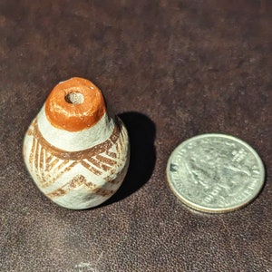 May include: A small, white ceramic pot with brown geometric designs. The pot has a round body and a small opening at the top. The pot is about the size of a US quarter.
