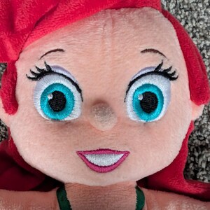 May include: A plush doll of a cartoon character with red hair, large turquoise eyes, and a smiling mouth. The doll is wearing a purple top with green straps. The doll's face is light pink.
