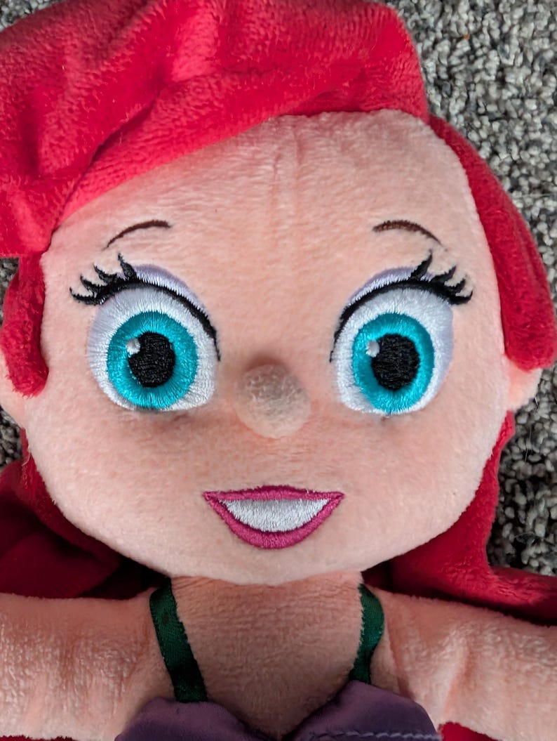 May include: A plush doll of a cartoon character with red hair, large turquoise eyes, and a smiling mouth. The doll is wearing a purple top with green straps. The doll's face is light pink.