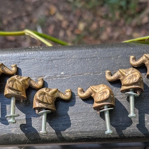 May include: A set of seven brass-toned cabinet knobs, each shaped like an elephant head with a trunk facing upward. Each knob features a metallic finish and includes a silver-colored metal bolt extending from the base for installation.